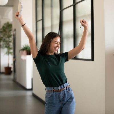 Emotional student girl within walls of institute celebrating successful exam pass, high school or college admission, european employee raised hands feels happy by salary growth, got promoted concept
